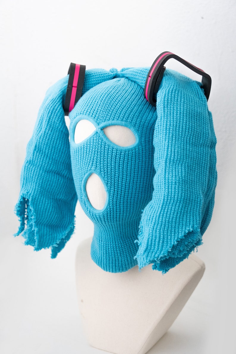 May include: A blue knitted balaclava with eye and mouth holes, featuring two long blue knitted strands with black and pink bands attached to the top.
