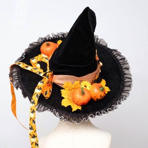 May include: A black velvet witch hat featuring a wide brim with a ruffled edge, decorated with orange pumpkins, yellow autumn leaves, a small yellow flower, and a long orange ribbon patterned with black silhouettes of flying witches.