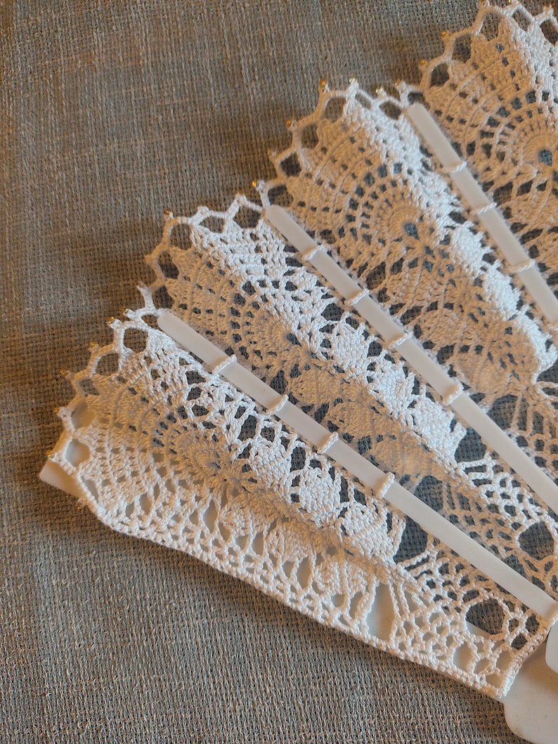 Puede incluir: Un abanico de crochet blanco con un delicado patr&oacute;n de encaje. El abanico est&aacute; plegado y tiene un marco de pl&aacute;stico blanco.