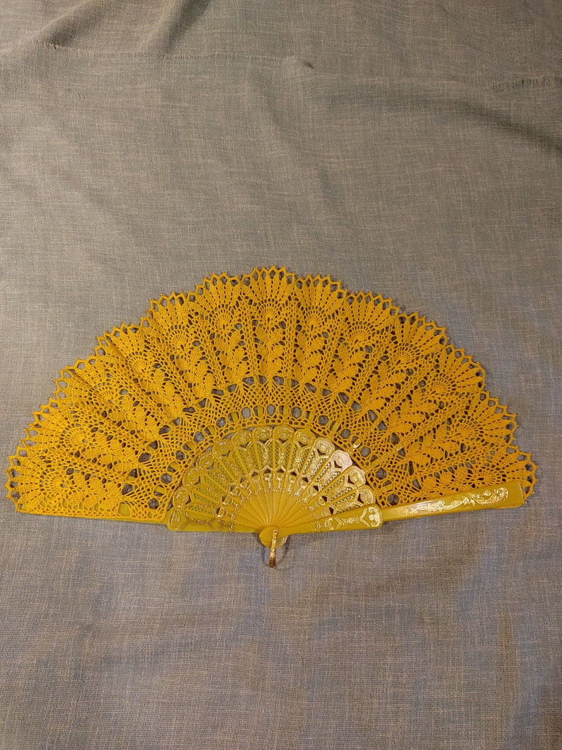 Puede incluir: Un abanico de crochet amarillo con un mango decorativo. El abanico est&aacute; cerrado y tiene un delicado patr&oacute;n de encaje.
