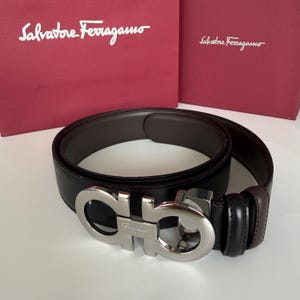 Könnte beinhalten: Ein schwarzer Ledergürtel mit einer silberfarbenen Gancini-Schnalle aus Metall, die das Salvatore Ferragamo-Logo trägt. Der Gürtel liegt vor einer roten Salvatore Ferragamo-Einkaufstasche und einer passenden roten Schachtel mit weißem Markennamen.