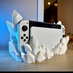 May include: A white Nintendo Switch console is displayed in a white, geometric crystal-shaped holder. The console has black buttons and a black screen border. The holder is made of white material and has a jagged, crystalline design.