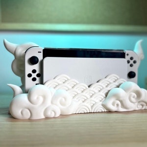 May include: A white Nintendo Switch console rests in a decorative white stand. The stand features a cloud-like design with curved edges and a wave pattern. The console's controllers are visible, and the overall aesthetic is minimalist.