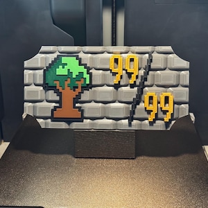 May include: A gray 3D-printed plaque with a brick wall texture featuring a pixelated green and brown tree icon on the left and the yellow pixelated numbers 99 over 99 on the right, separated by a black diagonal line, mounted on a black rectangular base.
