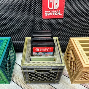 May include: A 3D-printed crate-style game cartridge holder, holding several black Nintendo Switch game cartridges. The holder is gold and sits on a wooden surface. A red Nintendo Switch logo is visible in the background.