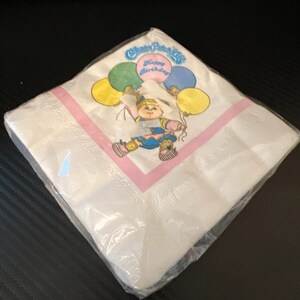May include: White paper napkin with a pink border. The napkin features a cartoon illustration of a girl holding balloons with the text "Cabbage Patch Kids Happy Birthday".