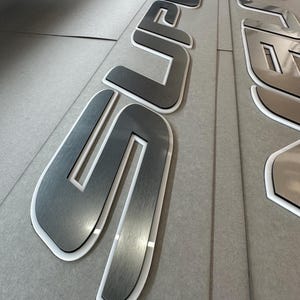 Puede incluir: Primer plano de letras met&aacute;licas dimensionales que deletrean "SUPER". Las letras tienen un acabado de metal cepillado con un borde blanco y est&aacute;n dispuestas sobre una superficie gris. Las letras son probablemente para se&ntilde;alizaci&oacute;n o exhibici&oacute;n.