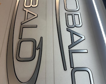 May include: Two metal signs with the word "ROBALO" in a stylized font. The letters are silver with a black outline. One sign has a curved design element, while the other is more linear. The signs are on a white background.