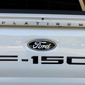 May include: The white tailgate of a Ford F-150 truck features a black Ford oval emblem centered above large, bold black F-150 lettering. Above the emblem, the word PLATINUM is spelled out in individual metallic silver letters on the tailgate.