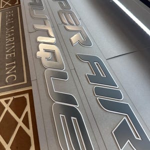 Puede incluir: Primer plano de letras de barco plateadas y grises. Las palabras "SUPER AIR NAUTIQUE" son visibles, con un contorno blanco. El texto "REAL MARINE INC" tambi&eacute;n es visible. Las letras est&aacute;n sobre una superficie gris.