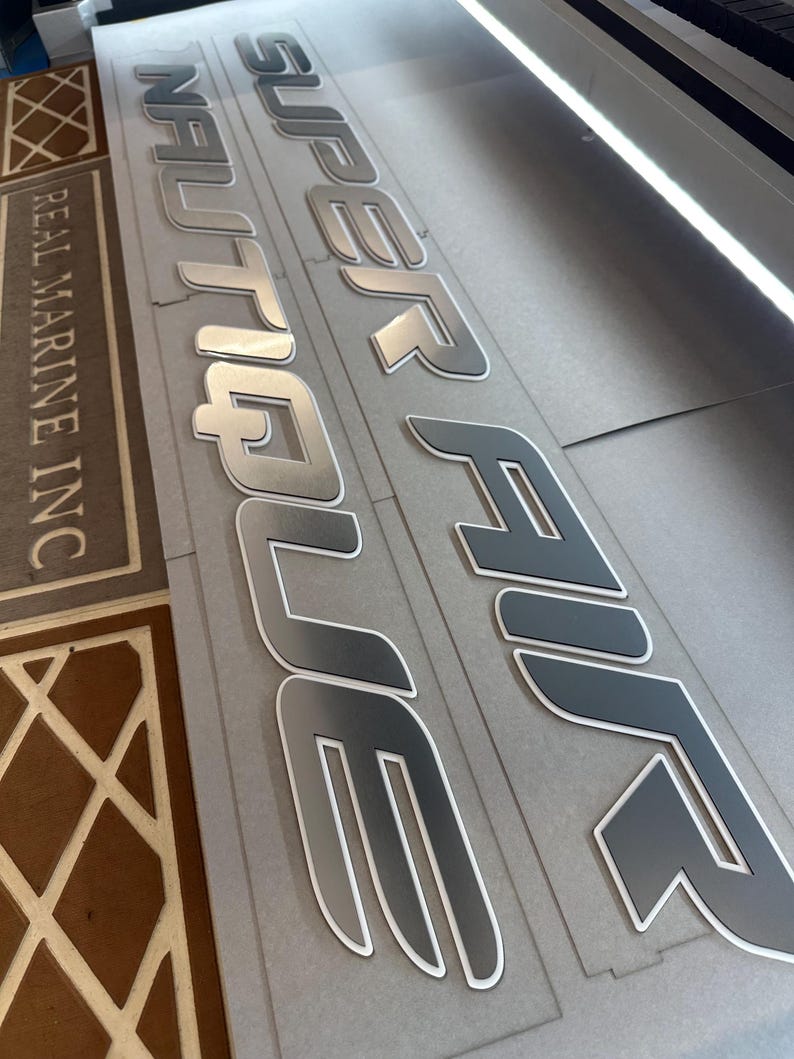 Puede incluir: Primer plano de letras de barco plateadas y grises. Las palabras "SUPER AIR NAUTIQUE" son visibles, con un contorno blanco. El texto "REAL MARINE INC" tambi&eacute;n es visible. Las letras est&aacute;n sobre una superficie gris.