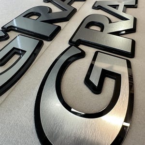 Puede incluir: Primer plano de varios emblemas automotrices negros y plateados. Las letras est&aacute;n en negrita, en una fuente sans-serif con un acabado plateado cepillado y un contorno negro. Los emblemas est&aacute;n dispuestos sobre una superficie de color claro.