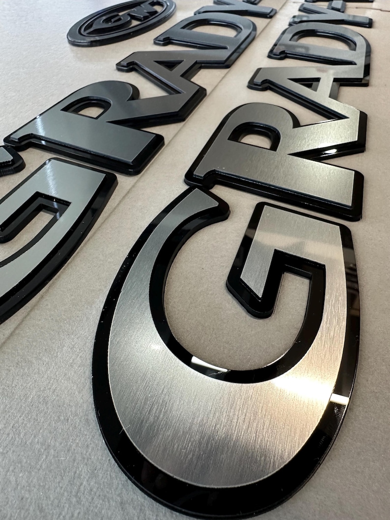 Puede incluir: Primer plano de varios emblemas automotrices negros y plateados. Las letras est&aacute;n en negrita, en una fuente sans-serif con un acabado plateado cepillado y un contorno negro. Los emblemas est&aacute;n dispuestos sobre una superficie de color claro.