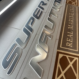 Puede incluir: Letras plateadas y grises para barcos con las palabras "SUPER AIR NAUTIQUE" y "REAL MARINE INC" impresas en una superficie gris. Las letras tienen un acabado met&aacute;lico con un contorno blanco.