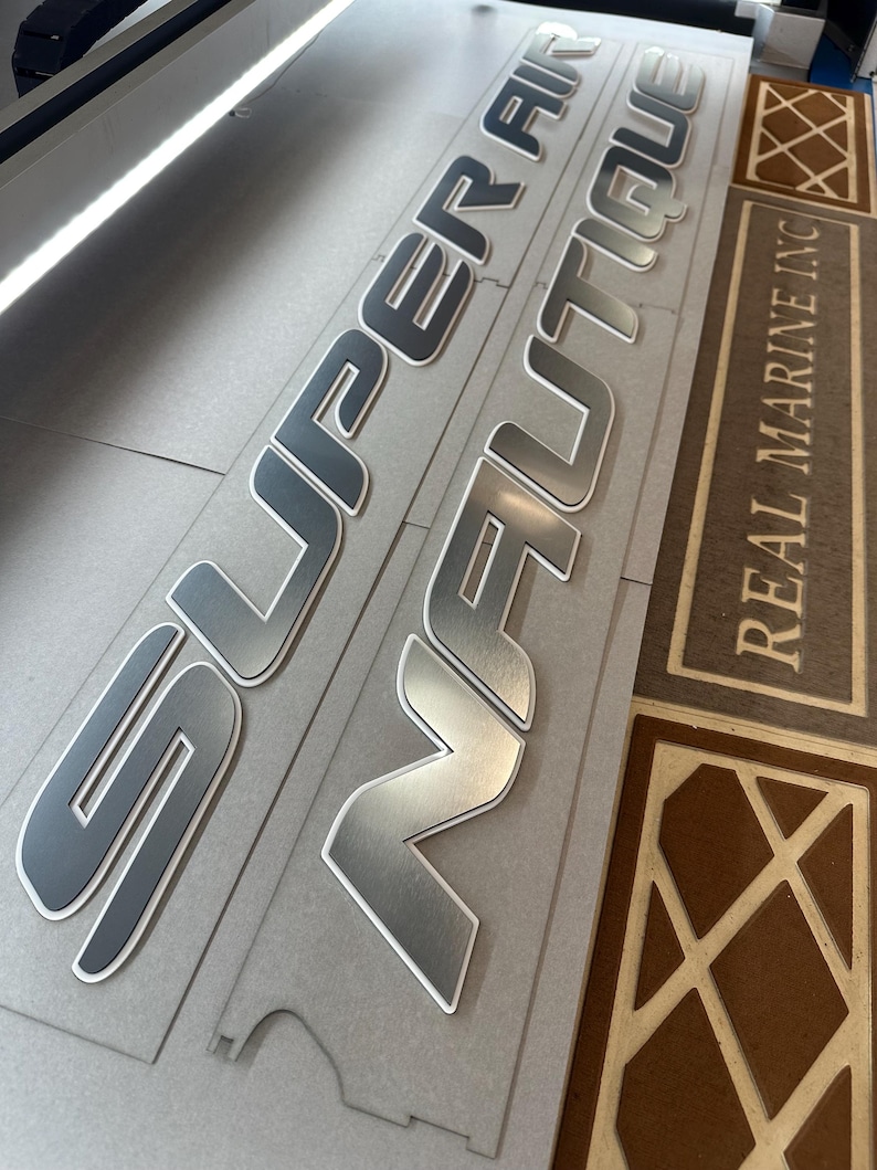 Puede incluir: Letras plateadas y grises para barcos con las palabras "SUPER AIR NAUTIQUE" y "REAL MARINE INC" impresas en una superficie gris. Las letras tienen un acabado met&aacute;lico con un contorno blanco.