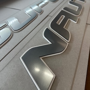 Puede incluir: Primer plano de letras met&aacute;licas plateadas con borde blanco, posiblemente para personalizaci&oacute;n de veh&iacute;culos. Las letras est&aacute;n sobre un fondo gris, con una superficie reflectante. La palabra "REVOLUTION" es parcialmente visible.