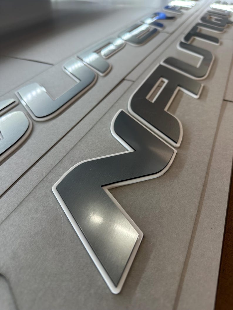 Puede incluir: Primer plano de letras met&aacute;licas plateadas con borde blanco, posiblemente para personalizaci&oacute;n de veh&iacute;culos. Las letras est&aacute;n sobre un fondo gris, con una superficie reflectante. La palabra "REVOLUTION" es parcialmente visible.