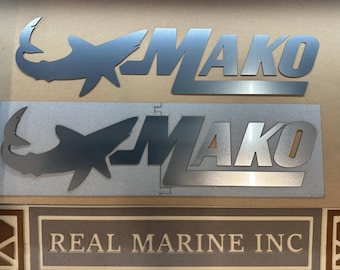 May include: Metal signs featuring the word "MAKO" with a shark silhouette. The top sign is a standalone design, while the bottom sign is mounted on a rectangular metal plate. The bottom of the image shows a sign that reads "REAL MARINE INC."