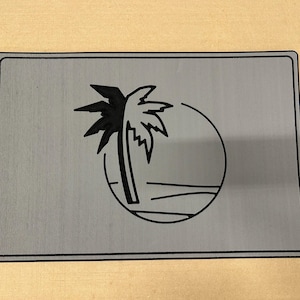 May include: A rectangular, grey metal sign with a black border. The sign features a black palm tree silhouette within a circle, with a line representing the horizon. The design evokes a tropical theme.