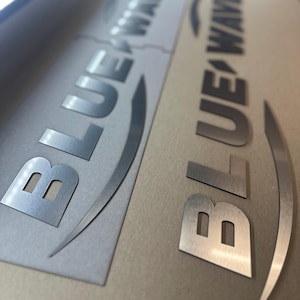 May include: Two metallic silver decals featuring the text BLUE WAVE in a bold, sans-serif font with a curved swoosh graphic underneath. The decals have a brushed metal finish and are displayed on a flat, light brown surface with a smooth texture.