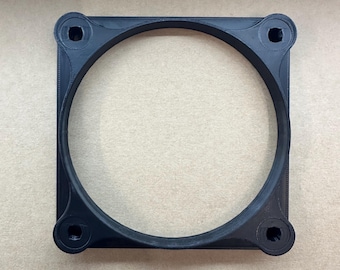 May include: A black square-shaped fan mounting bracket featuring a large circular opening in the centre and four reinforced circular corner mounts, each containing a small hole for screw attachment, constructed with a visible layered texture.