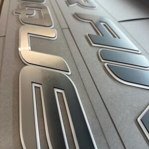 Puede incluir: Primer plano de letras met&aacute;licas plateadas con un contorno blanco. Las letras deletrean una palabra, posiblemente un nombre de marca o un identificador de producto. Las letras est&aacute;n sobre una superficie gris.