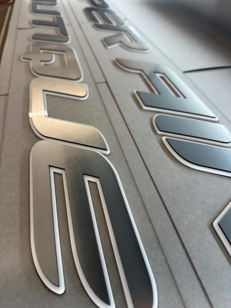 Puede incluir: Primer plano de letras met&aacute;licas plateadas con un contorno blanco. Las letras deletrean una palabra, posiblemente un nombre de marca o un identificador de producto. Las letras est&aacute;n sobre una superficie gris.