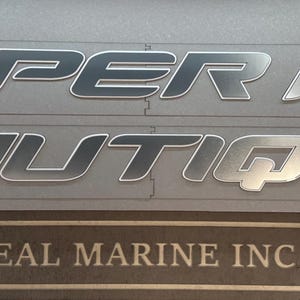 Puede incluir: Letras plateadas met&aacute;licas para barcos. Las palabras "SUPER AIR NAUTIQUE" se muestran en una fuente en negrita sin serifa. Debajo, un letrero rectangular que dice "REAL MARINE INC". El fondo es gris claro.