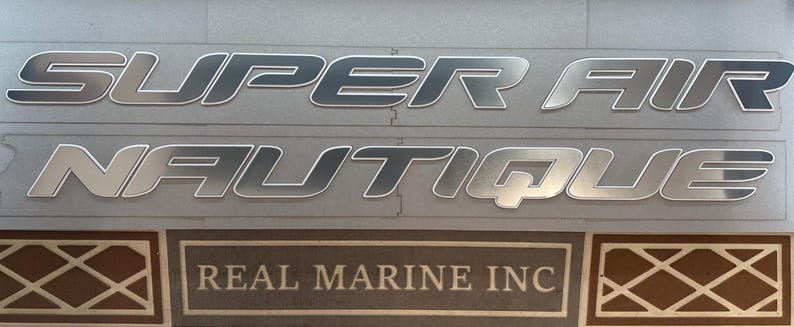 Puede incluir: Letras plateadas met&aacute;licas para barcos. Las palabras "SUPER AIR NAUTIQUE" se muestran en una fuente en negrita sin serifa. Debajo, un letrero rectangular que dice "REAL MARINE INC". El fondo es gris claro.
