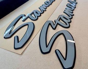 May include: Two gray and black vinyl decals with the word "Stamas" written in a cursive font.