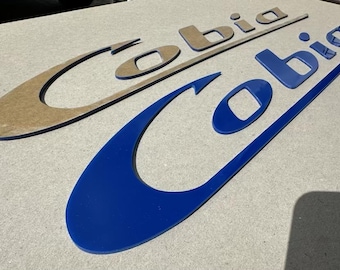 Könnte beinhalten: Zwei "Cobia"-Logos, eines in Braun und eines in Blau, werden auf einer hellgrauen Oberfläche angezeigt. Die Logos haben ein stilisiertes Design mit gebogenen Elementen und rechteckigen Ausschnitten. Das blaue Logo ist prominenter.