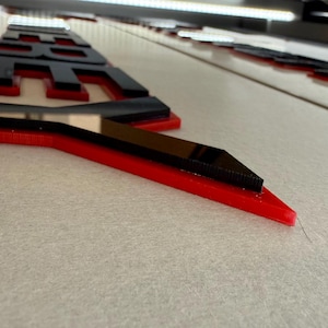 Puede incluir: Primer plano de un letrero rojo y negro con la palabra "RE" visible. Las letras son negras con un contorno rojo. El letrero est&aacute; sobre una superficie blanca.