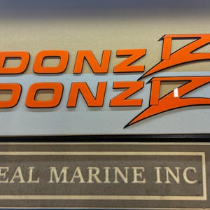 Puede incluir: Letrero con la palabra "DONZ" en letras de bloque naranja, con dise&ntilde;os estilizados de "Z" en naranja y negro. Debajo, el texto "REAL MARINE INC" se muestra en un letrero enmarcado de estilo vintage.