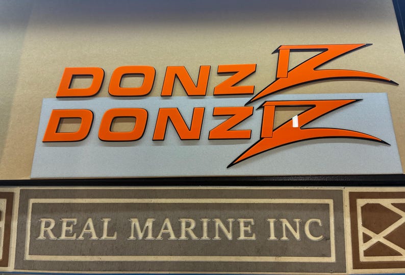 Puede incluir: Letrero con la palabra "DONZ" en letras de bloque naranja, con dise&ntilde;os estilizados de "Z" en naranja y negro. Debajo, el texto "REAL MARINE INC" se muestra en un letrero enmarcado de estilo vintage.