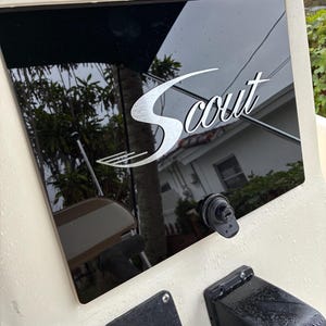 May include: A boat dashboard featuring the word "Scout" in a silver, stylized font. The dashboard is black and reflective, set within a cream-coloured frame. Other boat controls are visible.