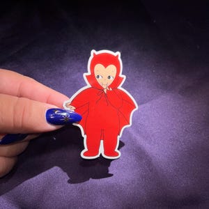 May include: A red devil-themed sticker with a cartoon illustration of a smiling character in a devil costume. The sticker has a white border and is held against a dark purple background.