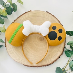 Può includere: Un paio di orecchie da topo marroni e bianche con un'impronta di zampa e un design a forma di osso. Le orecchie sono su uno sfondo di legno.