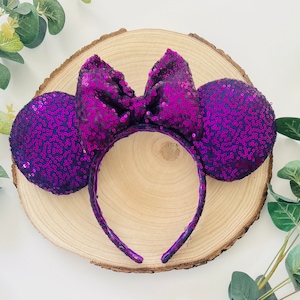 Op de afbeelding: Paarse Minnie Mouse oren met pailletten en een grote strik. De hoofdband ligt op een houten oppervlak.