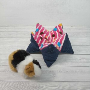 May include: A pink and blue patterned star-shaped bed for a small pet, such as a guinea pig or hamster. The bed is made of soft fabric and has a denim outer layer. A guinea pig is sitting next to the bed.