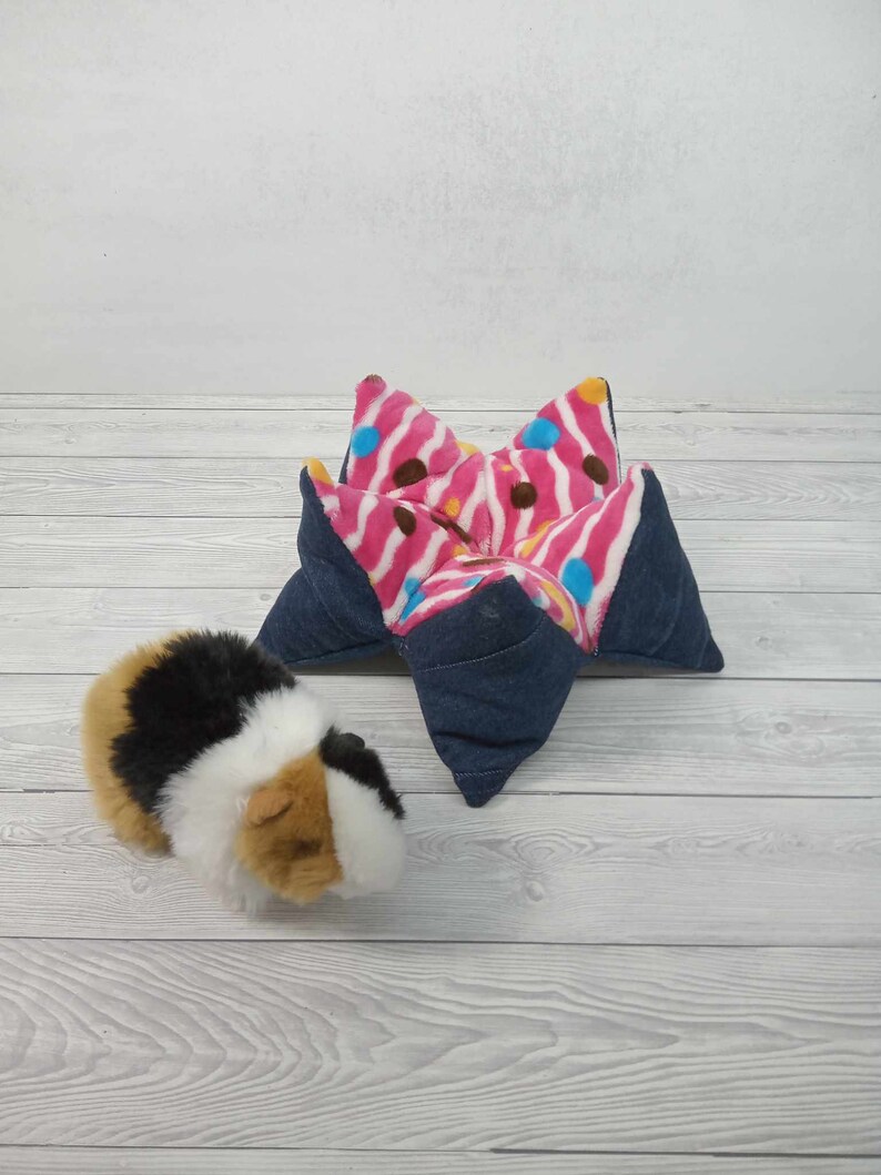 May include: A pink and blue patterned star-shaped bed for a small pet, such as a guinea pig or hamster. The bed is made of soft fabric and has a denim outer layer. A guinea pig is sitting next to the bed.
