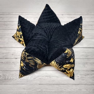 May include: A black and gold star-shaped floor cushion with a soft, plush texture. The cushion is made of two different fabrics, one with a black, textured surface and the other with a black and gold floral pattern.