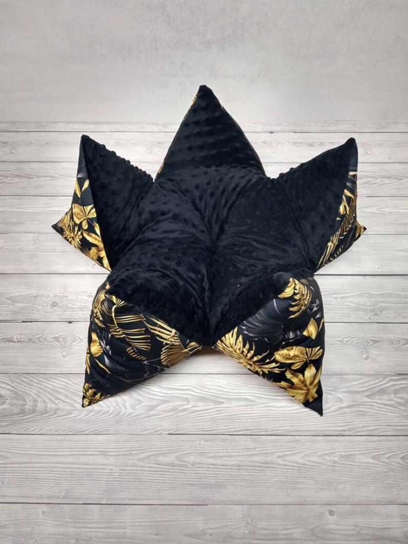 May include: A black and gold star-shaped floor cushion with a soft, plush texture. The cushion is made of two different fabrics, one with a black, textured surface and the other with a black and gold floral pattern.