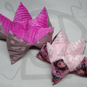 May include: Two pink star-shaped pillows with different patterns. The larger pillow has a pink and gray polka dot pattern, while the smaller pillow has a floral pattern.