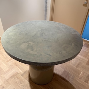 May include: A round, light grey concrete table with a beige pedestal base. The table is sitting on a light brown wood floor.