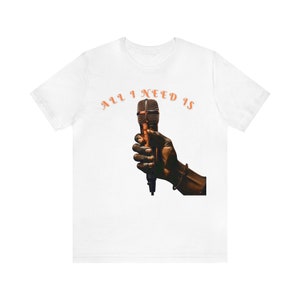 May include: White t-shirt featuring the phrase "ALL I NEED IS" in orange arched text above a graphic of a hand holding a bronze microphone. The hand and microphone are the focal point of the design.