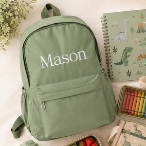 May include: A sage green backpack with the name Mason printed in white, a spiral notebook with dinosaur illustrations, a beige plush triceratops, a box of colorful crayons, a pencil case with dinosaur prints, a green water bottle, and a lunch container.