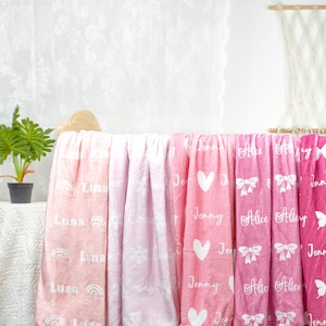 May include: A display of soft, personalized blankets in shades of pink. Each blanket features a name in white script, along with decorative elements like rainbows, hearts, bows, and butterflies. The blankets are draped over a wooden structure.