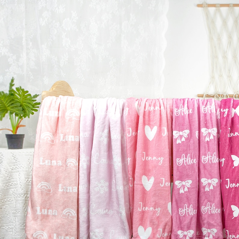 May include: A collection of soft fleece blankets in varying shades of pink, ranging from pale peach to deep magenta. Each blanket features a repeating white pattern of names like Luna, Laura, Jenny, and Alice, alongside icons of rainbows, snowflakes, hearts, bows, and butterflies.