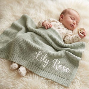Könnte beinhalten: Eine salbeigrüne Strickdecke für Babys mit dem in weißer Schreibschrift eingestickten Namen Lily Rose. Die Decke hat ein strukturiertes Strickmuster mit geripptem Rand und liegt über einem schlafenden Säugling in einem geblümten Strampler.