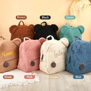 K&ouml;nnte beinhalten: Eine Auswahl an Rucks&auml;cken in Teddyb&auml;r-Form in verschiedenen Farben: Braun, Schwarz, Gr&uuml;n, Khaki, Rosa, Creme und Blau. Jeder Rucksack hat einen Rei&szlig;verschluss, einen kleinen runden Anh&auml;nger und gestickte Namen.